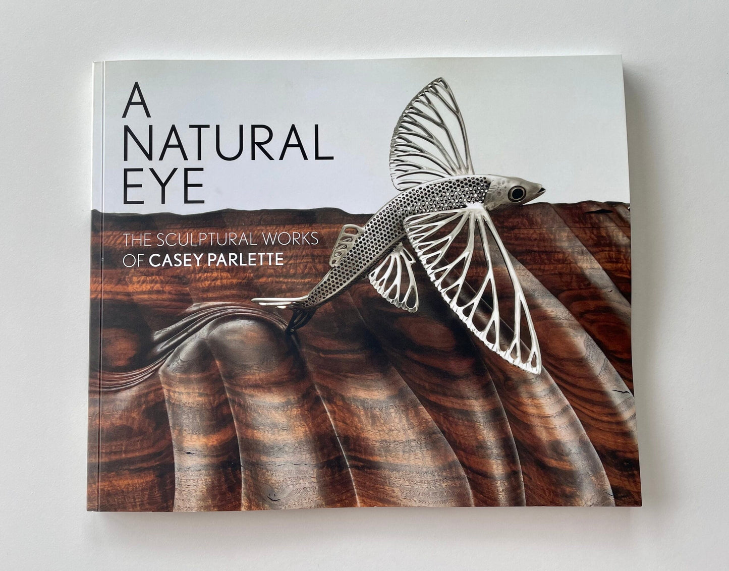 Cover of the book 'A Natural Eye: The Sculptural Works of Casey Parlette' featuring a sculpture of a flying fish on a wooden surface.